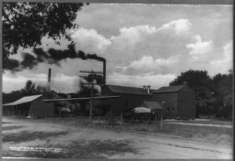 Black and white photograph of a cotton gin with smoking stacks, wagons drawn up in the yard in front.