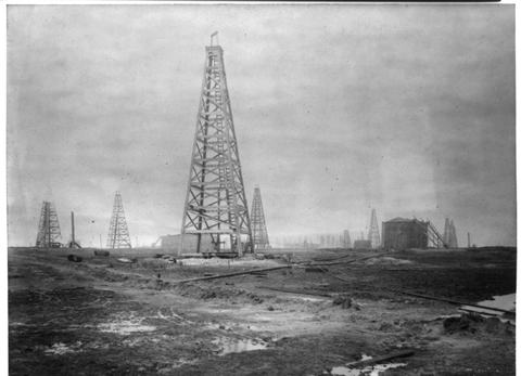 Black and white photograph of a tall wooden oil derrick on bare, muddy ground, with more derricks in the distance.