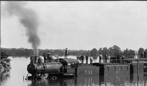 Black and white photograph of a steam locomotive and freight cars standing in floodwater, with men on the car roofs.